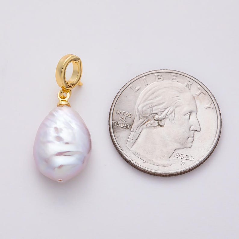 May include: A gold-colored earring pendant with a large, pear-shaped, iridescent, light purple pearl. The pendant is next to a U.S. quarter coin for size comparison. The coin shows the year 2022.