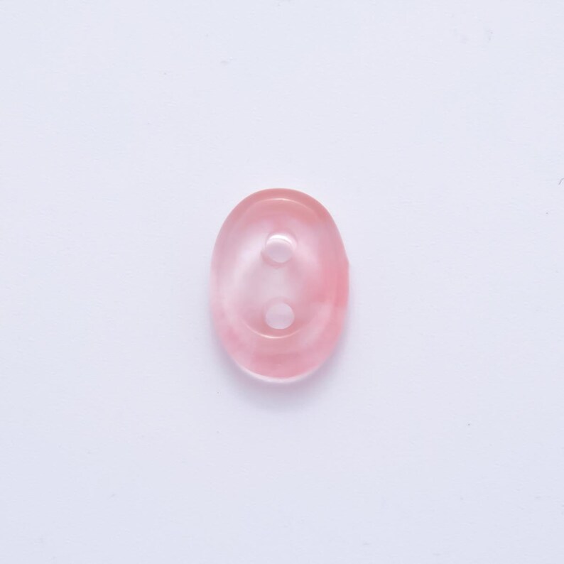 May include: Oval-shaped, translucent pink button with two holes. The button has a subtle, marbled appearance, with variations in the pink hue. The button is a sewing or craft supply.