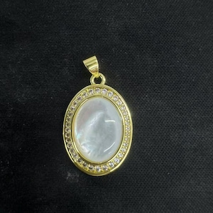 Puede incluir: Colgante ovalado dorado con una piedra central nacarada, rodeada por un anillo de pequeños cristales transparentes. El colgante tiene una argolla simple para una cadena. El fondo es una tela oscura y texturizada.