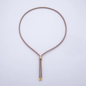 May include: A taupe-colored necklace with a circular design. The necklace features a gold-colored, rhinestone-studded centerpiece and two dangling cords with gold-colored tips. The necklace is displayed against a white background.