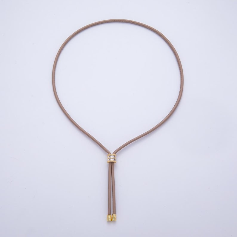 May include: A taupe-colored necklace with a circular design. The necklace features a gold-colored, rhinestone-studded centerpiece and two dangling cords with gold-colored tips. The necklace is displayed against a white background.