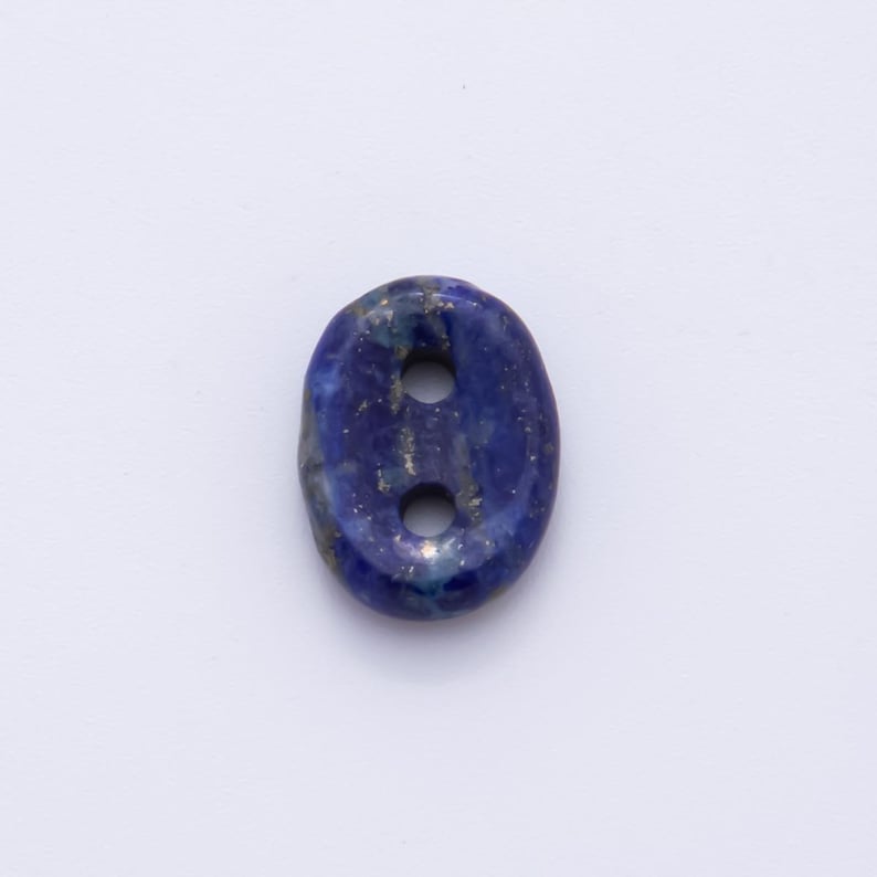 May include: Oval-shaped lapis lazuli button with two holes. The button is a deep blue color with flecks of gold throughout. The button is set against a white background.