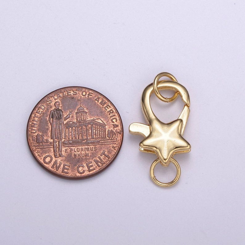 May include: Gold-toned metal star-shaped clasp with a ring for attaching to a necklace or bracelet. The clasp is shown next to a US penny for size comparison.