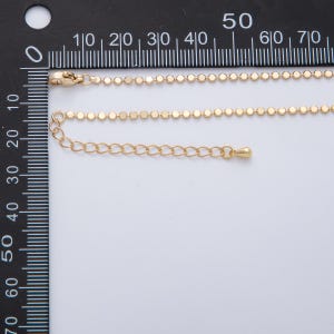 May include: Gold-colored necklace with a delicate chain of small, flat, round beads. The necklace includes a lobster clasp and an adjustable chain. The image is taken against a white background with a ruler in the background.