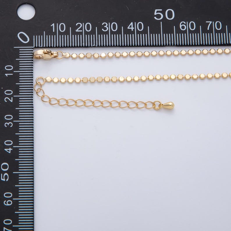May include: Gold-colored necklace with a delicate chain of small, flat, round beads. The necklace includes a lobster clasp and an adjustable chain. The image is taken against a white background with a ruler in the background.
