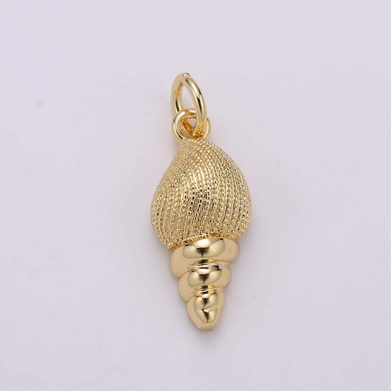 May include: A gold-coloured seashell charm with a textured surface and a spiral design. The charm has a loop at the top for attaching to a necklace or bracelet.