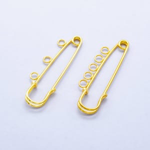May include: Two gold safety pins with multiple loops for attaching items. The pins are about 2 inches long.