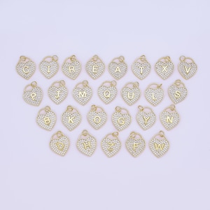 May include: A selection of gold-coloured heart-shaped pendants, each encrusted with small, clear crystals. Each pendant features a different letter of the alphabet in a gold tone. The pendants are arranged on a light purple background.