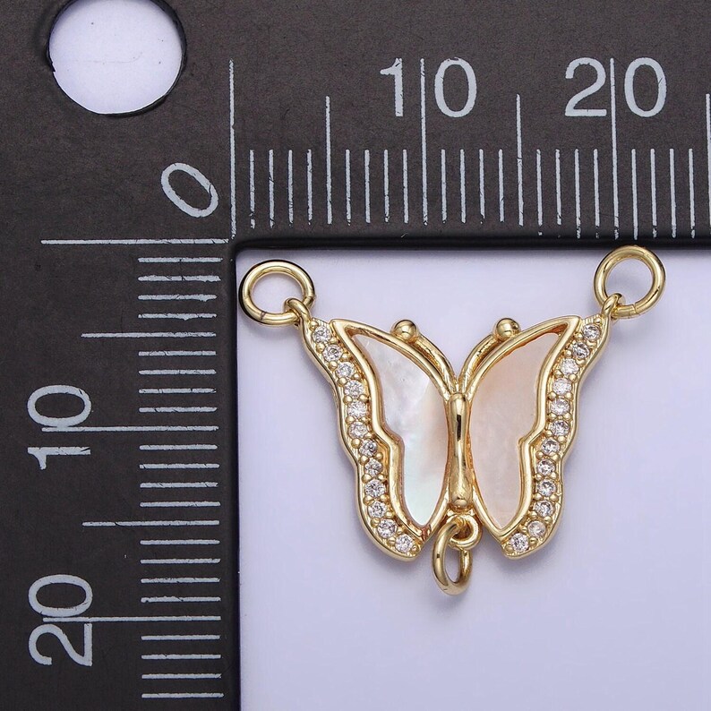 May include: Gold-tone butterfly charm with clear rhinestones and a white mother-of-pearl inlay. The charm has two jump rings for attaching to a necklace or bracelet.