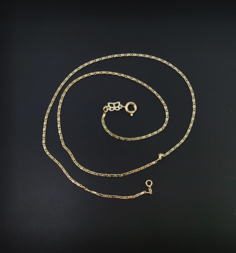 May include: A delicate gold-colored necklace with a small clasp and a decorative floral element. The chain is made of small, rectangular links. The necklace is displayed on a black background.