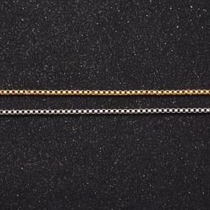 May include: Two chains, one gold and one silver, are laid out on a black textured surface. The chains are both box chains, with square links.