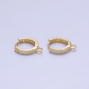 May include: A pair of gold-coloured hoop earrings, each featuring a row of small, clear gemstones. The earrings have a hinged closure and a small loop. They are displayed against a pale purple backdrop.