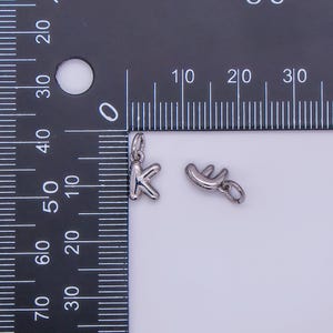 May include: Two small, silver-colored charms. One charm is the letter "K", and the other is a stylized shape. Both charms have a small loop at the top for attaching to a chain or bracelet. The charms are next to a ruler.