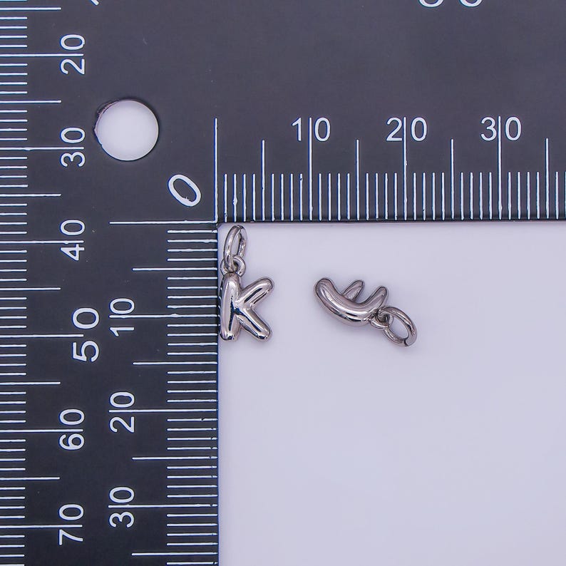 May include: Two small, silver-colored charms. One charm is the letter "K", and the other is a stylized shape. Both charms have a small loop at the top for attaching to a chain or bracelet. The charms are next to a ruler.