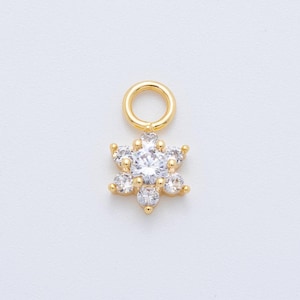 May include: A gold-colored charm with a flower design. The charm features a central, large, clear gemstone surrounded by six smaller clear gemstones. A small gold loop is at the top for attachment. The charm is likely for jewelry making.