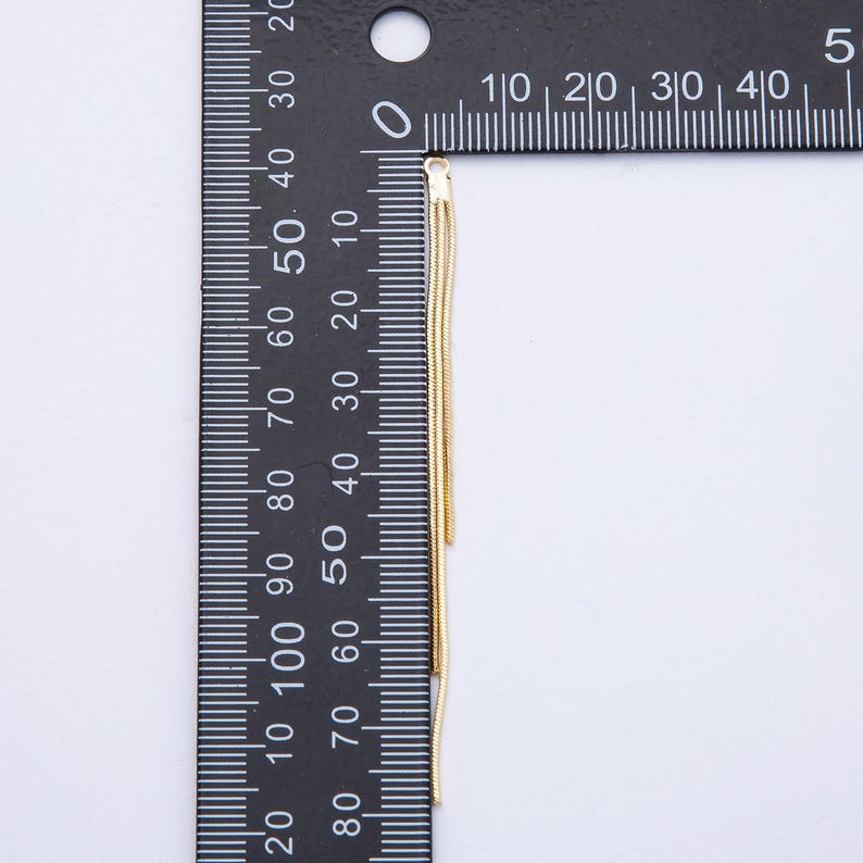 May include: A gold-colored, multi-strand chain, likely for jewelry, hangs vertically against a black ruler. The ruler displays centimeter measurements. The chain's delicate strands are visible, suggesting a lightweight design. The background is a plain white surface.
