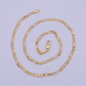 May include: A gold-colored Figaro chain necklace. The necklace features a pattern of three small circular links followed by one elongated oval link. The chain is displayed on a light purple background and has a lobster clasp closure.