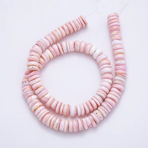 May include: A long strand of small, circular, disc-shaped beads in varying shades of soft pink and white. The beads are strung together on a thin white thread, displaying a smooth, polished texture with natural variations in color and thickness.