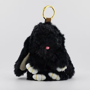 May include: A black plush bunny keychain with a gold-colored ring and brown strap. The bunny has a pink nose, white paws with gold stitching, and cream-colored inner ears. The plush toy is fluffy and soft.