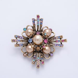 May include: A gold-toned brooch with a floral design. The brooch features faux pearls and colorful rhinestones in shades of pink, blue, and yellow. The brooch is set against a white background.