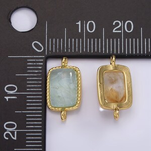 May include: Two gold-toned metal charms with square-shaped frames and clear stones. The charm on the left has a light blue stone, and the charm on the right has a light brown stone. Both charms have two loops on the top for attaching to a necklace or bracelet.