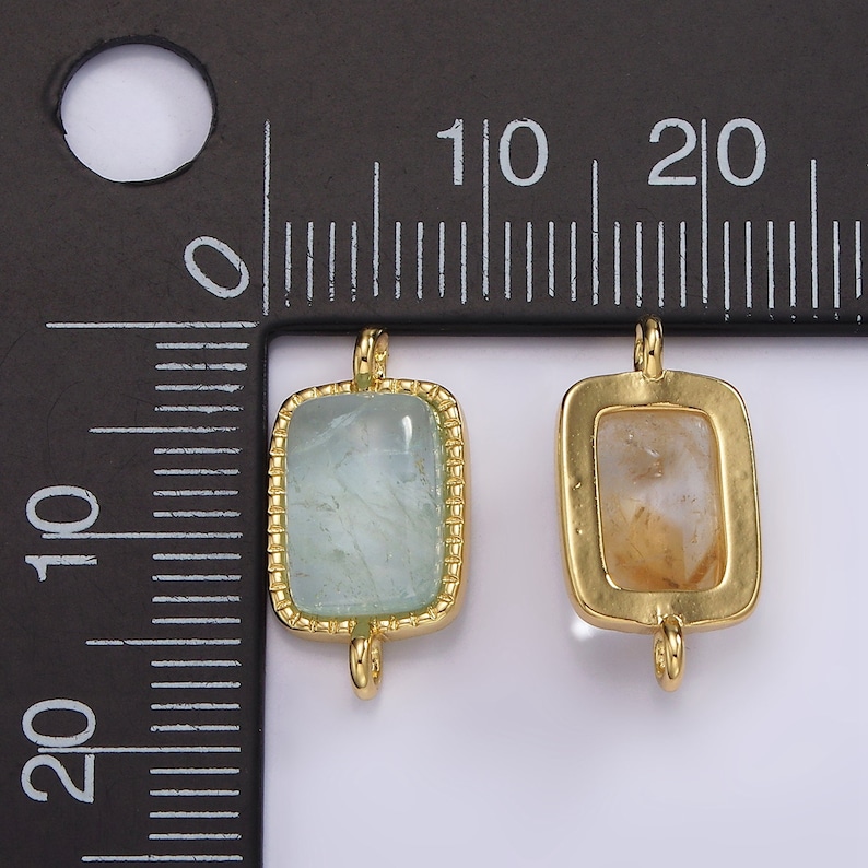 May include: Two gold-toned metal charms with square-shaped frames and clear stones. The charm on the left has a light blue stone, and the charm on the right has a light brown stone. Both charms have two loops on the top for attaching to a necklace or bracelet.