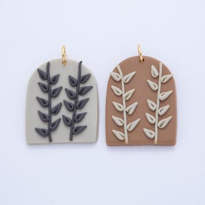 May include: Two clay pendants with a botanical design. One is light grey with dark grey leaves, the other is brown with off-white leaves. Each has a gold-coloured loop at the top for hanging.