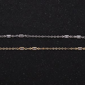 May include: Two delicate chains with rectangular clear stones, one silver and one gold, on a black background.
