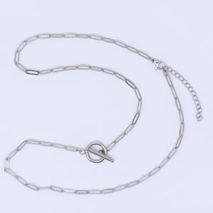 Könnte beinhalten: Eine silberfarbene Halskette mit einem Design aus ovalen Gliedern. Die Halskette hat einen Knebelverschluss. Die Kette wird vor einem weißen Hintergrund gezeigt.