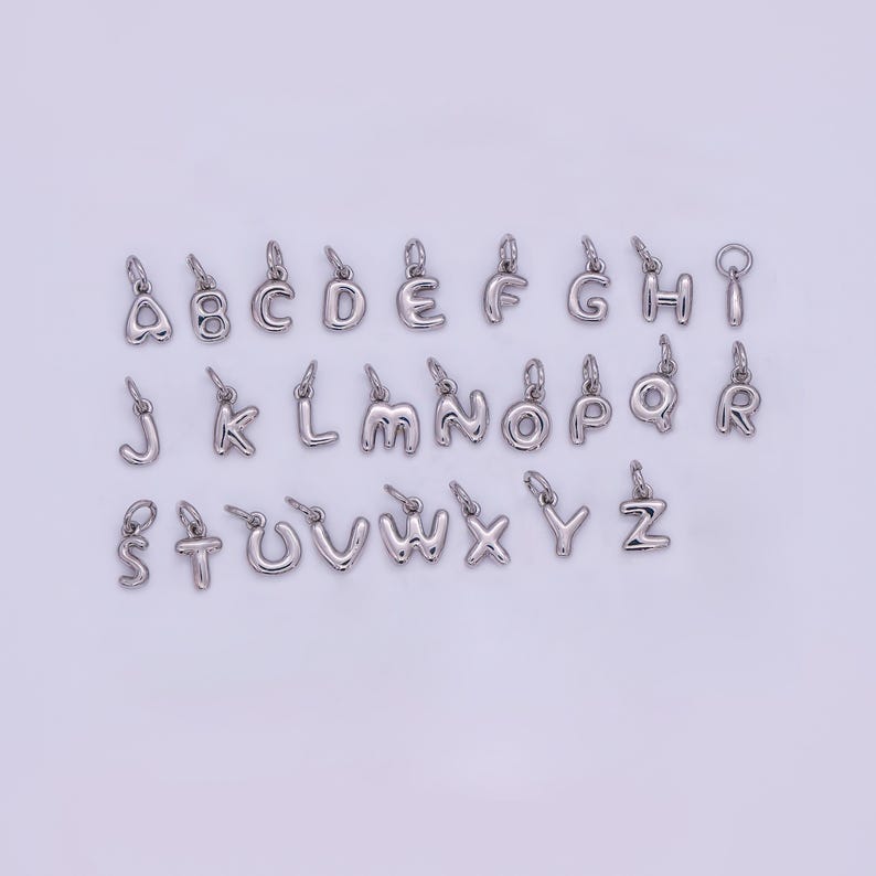 May include: A collection of silver-colored alphabet charms, each with a small loop for attaching to jewelry. The letters A through Z are displayed, showcasing a variety of fonts and styles. These charms are ideal for personalized necklaces or bracelets.