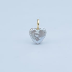 May include: A heart-shaped pendant featuring a lustrous, off-white pearl. The pearl is suspended from a small, gold-coloured bail. The pendant is set against a plain, light background.