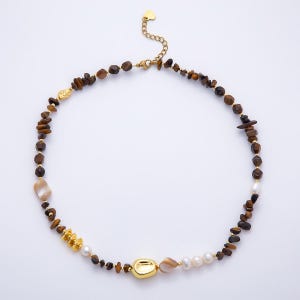 Puede incluir: Un collar con fragmentos de piedra ojo de tigre marrón, perlas de agua dulce blancas y detalles de metal dorado. La pieza incluye una cuenta central dorada irregular, espaciadores dorados más pequeños y una cadena extensora con un pequeño dije de corazón.