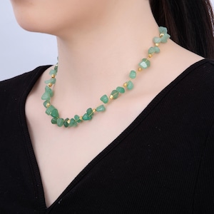 May include: A delicate necklace featuring small, irregular green gemstones strung on a gold-colored wire. The necklace has a simple design with small gold beads interspersed between the green stones. The necklace is worn against a black top.