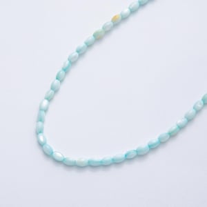 Puede incluir: Un collar o pulsera de cuentas ovaladas de color azul claro. Las cuentas están ensartadas, formando una línea curva. Las cuentas tienen un brillo nacarado y están colocadas sobre un fondo blanco.
