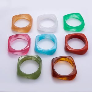 May include: Nine square-shaped resin rings in various colors, including orange, white, green, pink, blue, and brown. The rings have a smooth, glossy finish and a rounded inner edge. The rings are displayed on a white surface.