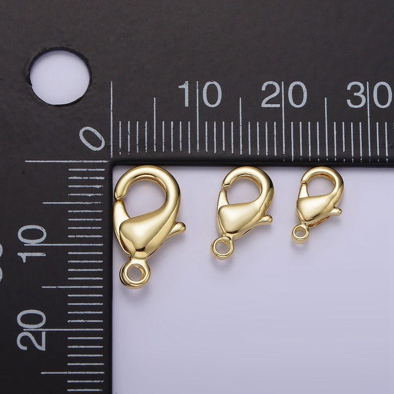 May include: Three gold-toned lobster clasps of different sizes, measured against a ruler in centimetres.