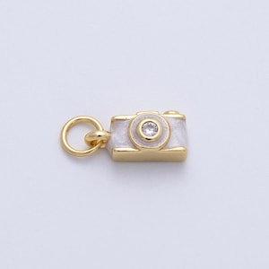May include: A gold-colored camera charm with a white enamel finish. The camera has a small, clear gemstone in the center of the lens and a gold loop for attaching to a necklace or bracelet. Jewelry accessory.