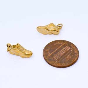 May include: Two gold-colored sneaker charms and a penny are displayed against a white background. The charms are detailed miniature replicas of athletic shoes. The penny is a copper-colored coin with the image of Abraham Lincoln.