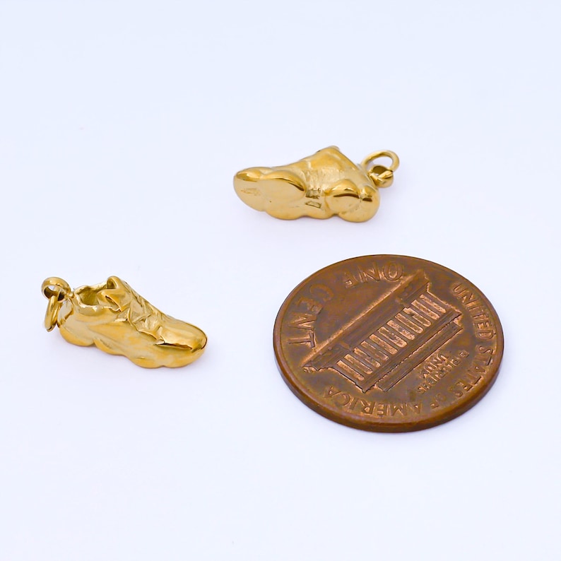 May include: Two gold-colored sneaker charms and a penny are displayed against a white background. The charms are detailed miniature replicas of athletic shoes. The penny is a copper-colored coin with the image of Abraham Lincoln.