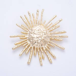 Pode incluir: Um broche dourado em forma de raio de sol, com um centro texturizado e pontas radiantes. O broche tem um acabamento metálico brilhante e é provavelmente feito de metal. Este acessório pode ser usado para embelezar roupas ou acessórios.
