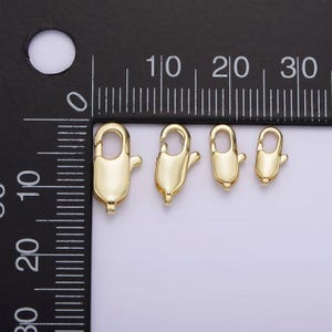 May include: Four gold-toned lobster clasps of different sizes, measured in centimeters on a black ruler.
