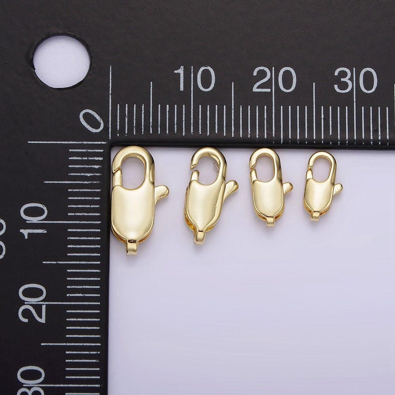 May include: Four gold-toned lobster clasps of different sizes, measured in centimeters on a black ruler.