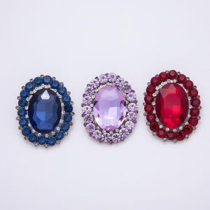 May include: Three oval-shaped brooches with large, faceted gemstones. The brooches are in shades of blue, lavender, and red, each surrounded by smaller, matching stones. The brooches are set against a white background.