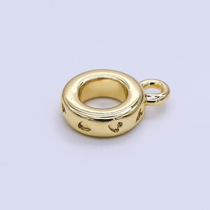 May include: A gold-colored charm with a circular design and a small loop. The charm has a series of heart-shaped cutouts around the outer edge. The charm is made of a shiny, reflective metal.