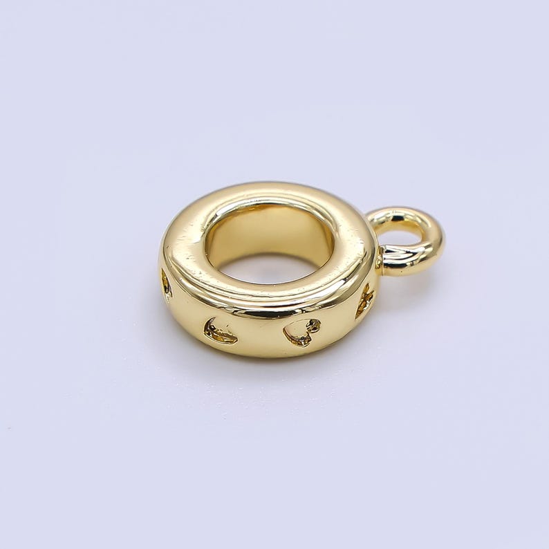 May include: A gold-colored charm with a circular design and a small loop. The charm has a series of heart-shaped cutouts around the outer edge. The charm is made of a shiny, reflective metal.