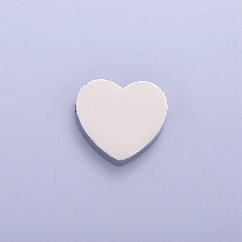 Puede incluir: Un charm de coraz&oacute;n plateado con una superficie lisa y blanca. El charm es perfecto para agregar un toque de amor a los proyectos de joyer&iacute;a.