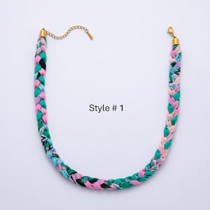 May include: Braided fabric necklace in shades of pink, green, and black with gold-colored chain and clasp. The necklace is labeled "Style # 1". The braided design adds a unique texture to this fashion accessory.