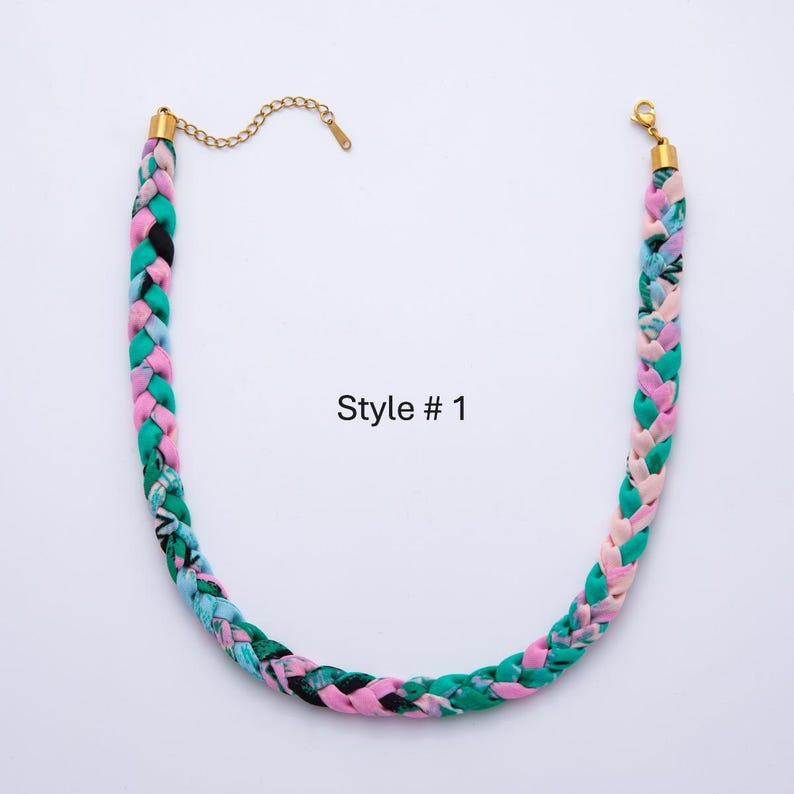 May include: Braided fabric necklace in shades of pink, green, and black with gold-colored chain and clasp. The necklace is labeled "Style # 1". The braided design adds a unique texture to this fashion accessory.