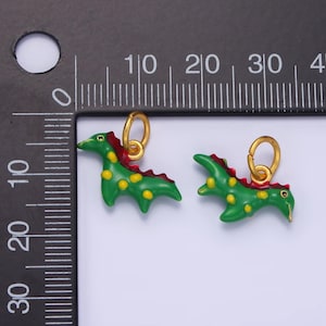 May include: Two green dinosaur charms with gold accents. The dinosaurs have red accents on their backs and yellow spots. The charms are hanging from gold jump rings.
