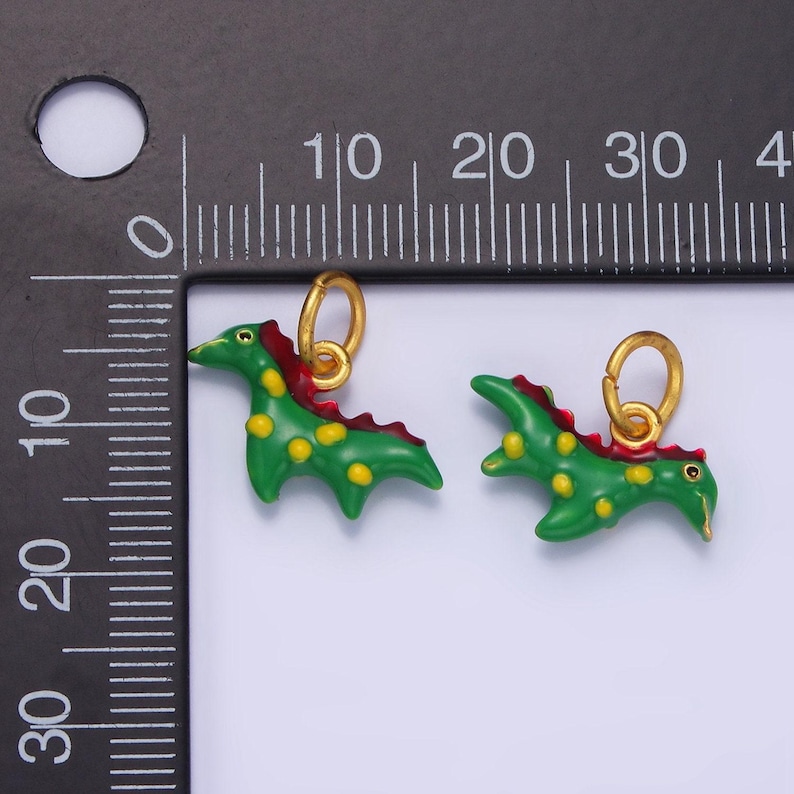 May include: Two green dinosaur charms with gold accents. The dinosaurs have red accents on their backs and yellow spots. The charms are hanging from gold jump rings.